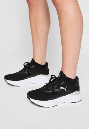 Beine, die schwarze Puma Sportschuhe mit weißen Sohlen auf einem schlichten hellen Hintergrund tragen.