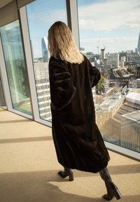 Breedide juustega naine, kes kannab pikka tumedat karvkattega mantlit ja musti saapaid, kõnnib klaasist seintega hoones, kust avaneb vaade linna siluetile päikesepaistelisel päeval.