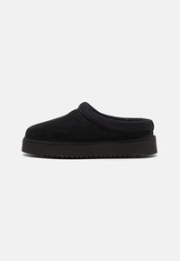 Zapato negro de ante tipo slip-on con punta redondeada, que cuenta con una parte superior acolchada y una suela de goma estriada. Textura suave, diseño minimalista.