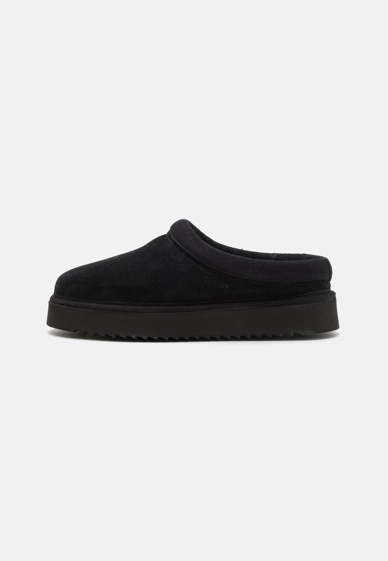 Zapato negro de ante tipo slip-on con punta redondeada, que cuenta con una parte superior acolchada y una suela de goma estriada. Textura suave, diseño minimalista.