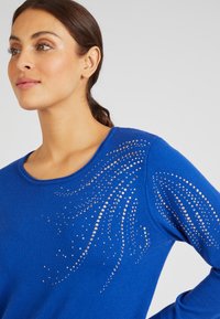 Blauwe gebreide trui met een ronde halslijn en decoratieve gouden rhinestones in een draaiend patroon op de linker schouder.