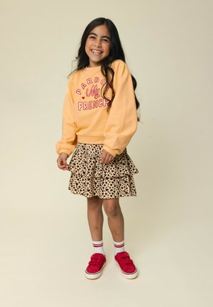 Lachend meisje met lang donker haar, gekleed in een gele trui, een rok met luipaardprint, witte sokken en rode sneakers, staand voor een beige achtergrond.