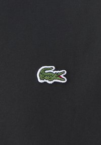 Juodas audinys su mažuoju siuvinėtu žaliu krokodilo logotipu, turinčiu baltą kontūrą ir raudoną akcentą ant burnos.