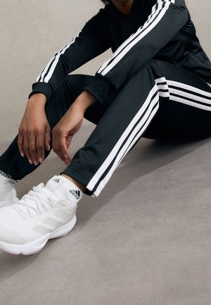 Osoba siedząca na podłodze, ubrana w czarny dres z białymi paskami oraz białe sneakersy Adidas z logo na skarpetkach i butach.