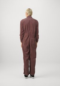Mono de pana burdeos con cuello, mangas largas, cintura elástica y bolsillo trasero. Tejido texturizado con líneas verticales.