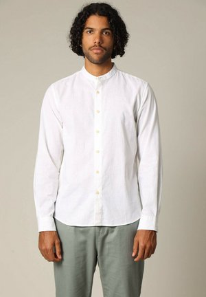 Chemise blanche à manches longues en lin avec un col mandarin, comportant une patte de boutonnage sur le devant et des boutons beige clair. Associée à un pantalon vert clair.