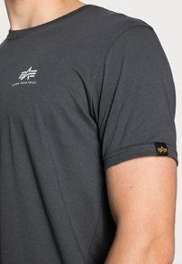 Dunkelgraues T-Shirt aus Baumwolle mit einem weißen Logo auf der Brust und einem kleinen schwarzen Etikett mit gelbem Akzent am Ärmel.