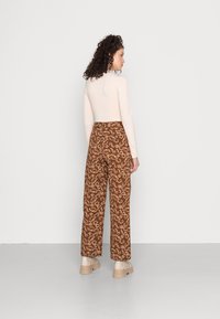 Camisola de manga longa bege combinada com calças largas castanhas com um padrão paisley em creme. A modelo está de pé em um ambiente neutro, usando sapatos chunky bege.