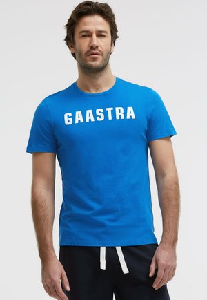 Man wearing blue short-sleeve t-shirt with "GAASTRA" text across the chest and black pants with white drawstrings standing against a plain background.