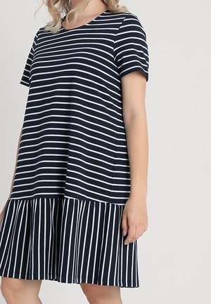 Vrouw die een marineblauwe jurk draagt met witte horizontale strepen op het bovenstuk en verticale strepen op de geplooide rok, staand tegen een effen achtergrond.