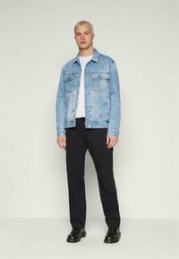 Heledad sinine teksajakk kahe rinnataskuga, valge T-särgi peal, tumedate pükstega ja mustade kingadega. Mudel seisab püsti.