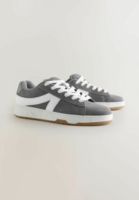 Une paire de baskets en daim gris avec des lacets blancs et une bande latérale blanche, posée sur un fond neutre clair.
