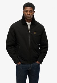 Veste noire zippée avec col en velours côtelé, poches latérales et un petit patch logo. Portée par-dessus un pull gris clair et un jean en denim foncé.