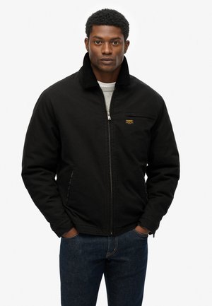 Schwarze Zip-Jacke mit Cordkragen, Seitentaschen und einem kleinen Logopatch. Tragen über einem hellgrauen Pullover und dunklen Jeans.
