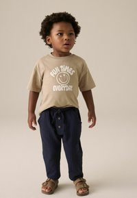 Beige T-Shirt mit weißem Text und Smiley-Gesicht; dunkelblaue gestreifte Hose mit Kordelzug und Holzknöpfen; braune Sandalen.