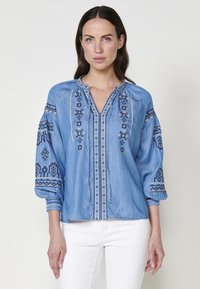 Blå chambrayskjorta med broderade detaljer på framsidan och ärmarna, med V-ringning och elastiska ärmslut.