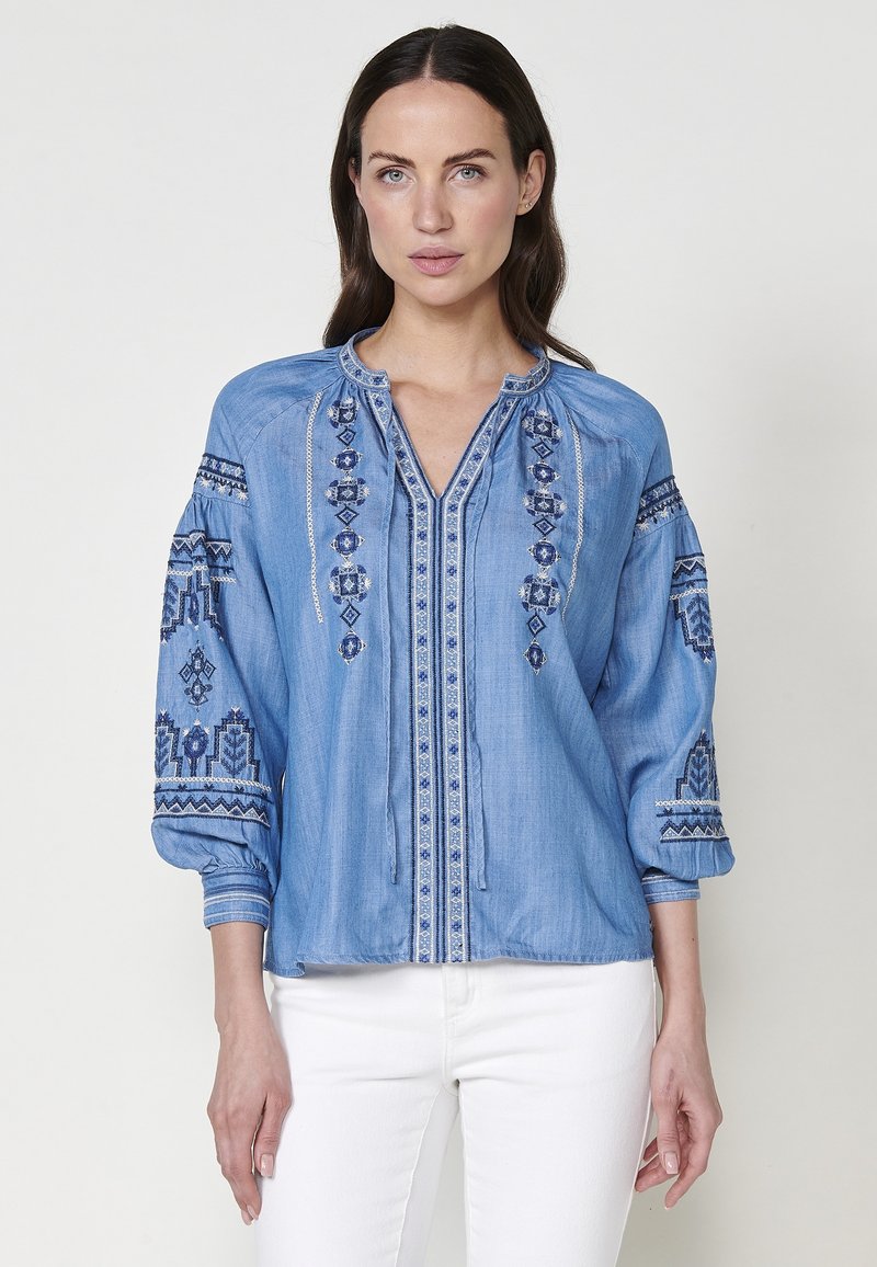 Blå chambrayskjorta med broderade detaljer på framsidan och ärmarna, med V-ringning och elastiska ärmslut.