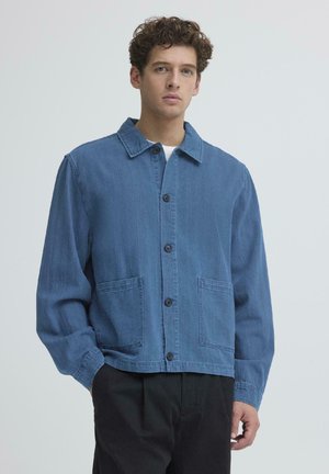 Junger Mann trägt eine blaue Jacke mit Knöpfen und zwei Vordertaschen sowie schwarze Hose, steht vor einem einfarbigen hellen Hintergrund.