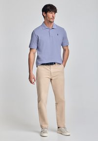 Polo shirt viola chiaro con colletto, indossato con chinos beige e sneakers bianche; presenta un logo ricamato sul petto, tessuto morbido.