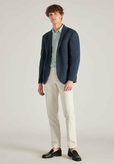 Donkerblauwe blazer over een lichtblauwe polo, gecombineerd met crèmekleurige broek en zwarte loafers. Herenigde texturen, op maat gemaakte pasvorm en gestructureerd ontwerp.