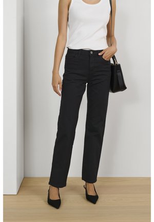 Frau trägt schwarze gerade geschnittene Jeans, weißes ärmelloses Oberteil, schwarze spitz zulaufende High Heels und hält eine schwarze Handtasche auf einem Holzboden.