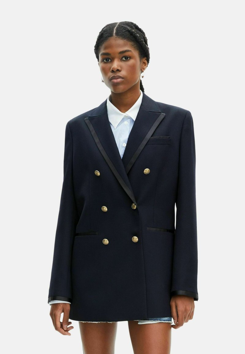 Blazer double petto blu navy con bottoni dorati, dettagli in raso sul bavero e due tasche frontali. Indossato sopra una camicia con colletto.