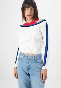 Hvit ribbet kortermet genser med off-the-shoulder design, med blå og svarte stripe detaljer langs ermene. Kombinert med lyseblå jeans.