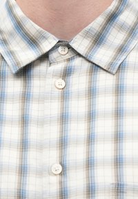 Nærbilde av en person som har på seg en hvit rutete skjorte med blå og brune striper, knyttet igjen ved kragen og midtknappen.