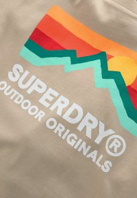 Bežová látka s multikolorovou grafikou hor, slunce a textem "SUPERDRY® OUTDOOR ORIGINALS" v tučné bílé.