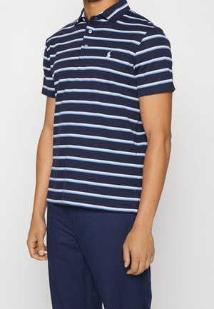 Mann trägt ein marineblau-hellblau gestreiftes kurzärmeliges Poloshirt mit Kragen und marineblaue Hose, steht vor einem schlichten Hintergrund.