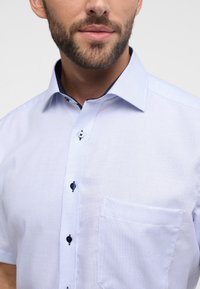 Hellblau karierte Kurzarmbluse mit Button-Down-Kragen, ausgestattet mit dunklen Knöpfen und einer Brusttasche. Glatte Stofftextur.