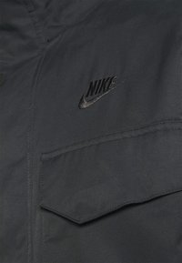Melns žakets ar teksturētu audumu, ar kabatu ar atloku un melnu izšūtu Nike logo krūtīs.