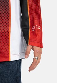 Sarkans un melns garām piedurknēm krekls ar punktu rakstu, baltajiem akcentiem un oranžu svītru. Uz piedurknes ir logotips, kā arī redzamas šuvju detaļas.