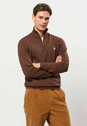 Jeune homme aux cheveux bruns portant un pull marron zippé et un pantalon en velours côtelé moutarde, debout les bras croisés devant un fond uni.