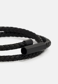 Mustvalge punutud nahkjuhe, mille mõlemas otsas on silindriline must metallist riistvara. Tekstuur on sile ja kompleksne, omades tihedat koet.