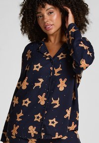 Marineblauwe pyjama bovenstuk van gladde stof, met patronen van peperkoekmannetjes, rendieren en sterren, met knoopsluiting en lange mouwen.