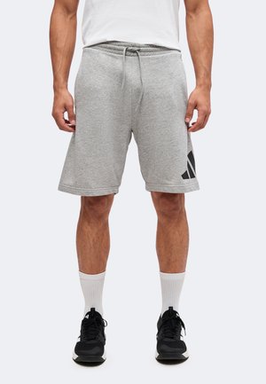 Mann trägt hellgraue Shorts mit Kordelzug, weiße Kniestrümpfe, schwarze Sportschuhe und ein weißes T-Shirt, steht vor neutralem Hintergrund.