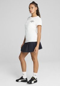 Witte katoenen t-shirt met zwart Puma-logo, gecombineerd met een marineblauwe geplooide rok en zwarte Puma sneakers, die een sportieve uitstraling en casual stijl laten zien.
