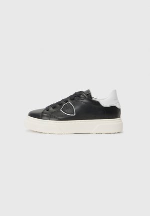 Black leather sneaker with a white rear accent, round toe, thick white sole, and decorative stitching. Features black laces and a logo detail.
