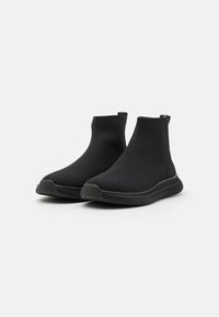 Ein Paar schwarze, gestrickte, knöchelhohe Slip-On-Sneaker mit strukturierten Sohlen auf einem weißen Hintergrund.