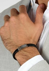 Svart flätad läderarmband med silverfästen och en texturerad rektangulär lås som har en logotyp. Bärs på en hand mot en ljus bakgrund.