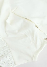 Vêtement en coton blanc avec une bordure en dentelle texturée en bas, doté de boutons-pression sur un côté pour la fermeture. Tissu doux avec une finition lisse.