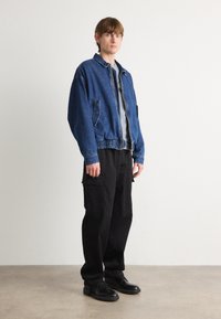 Veste en jean avec une fermeture éclair, deux poches avant et une taille élastique, portée sur une chemise grise, associée à un pantalon cargo noir ample et des chaussures noires.