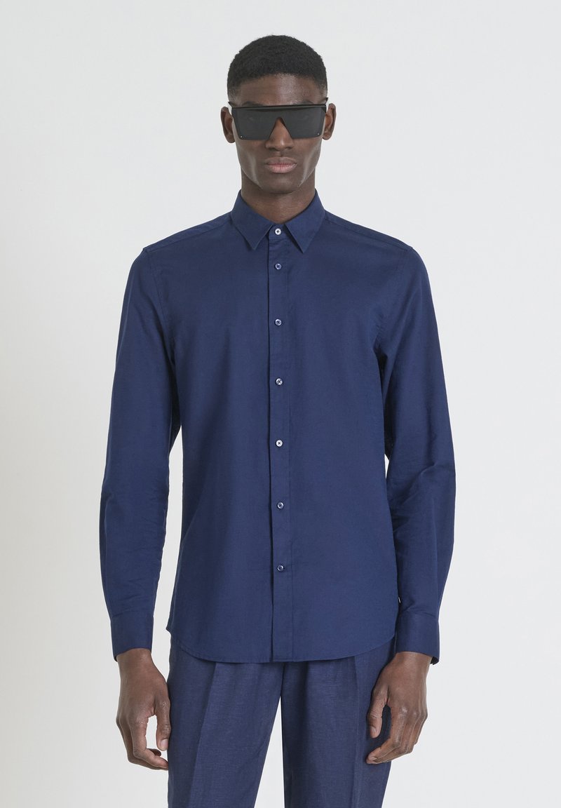 Camicia a maniche lunghe blu navy con colletto button-down, caratterizzata da una texture liscia e una vestibilità dritta, abbinata a pantaloni scuri e occhiali da sole.