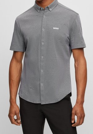 Man draagt een grijs overhemd met korte mouwen en knopen met het "BOSS"-logo op de borst, gecombineerd met een zwarte broek, tegen een effen grijze achtergrond.