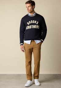 Marineblauer Pullover mit "BROOKS BROTHERS" in Creme, kombiniert mit einem hellblau gestreiften Hemd und beigen Hosen, ergänzt durch weiße Sneakers.