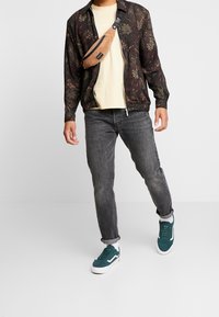 Man in zwarte paisley-patroon zip-jas, crème t-shirt, grijze opgerolde jeans, teal sneakers en een bruine corduroy schoudertas.