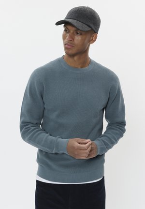 Sudadera gris de punto waffle con cuello redondo y puños acanalados, superpuesta a una camisa blanca, combinada con una gorra de fieltro oscura.