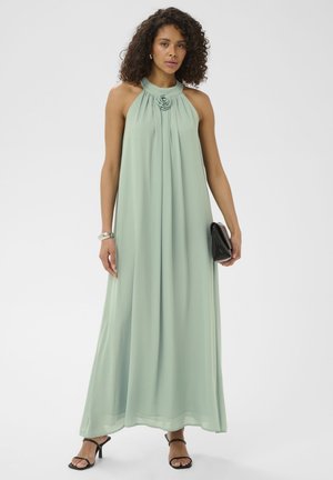 Kobieta o kręconych włosach w jasnozielonej, długiej do ziemi sukience bez rękawów, czarnych sandałach, trzymająca czarną kopertówkę.