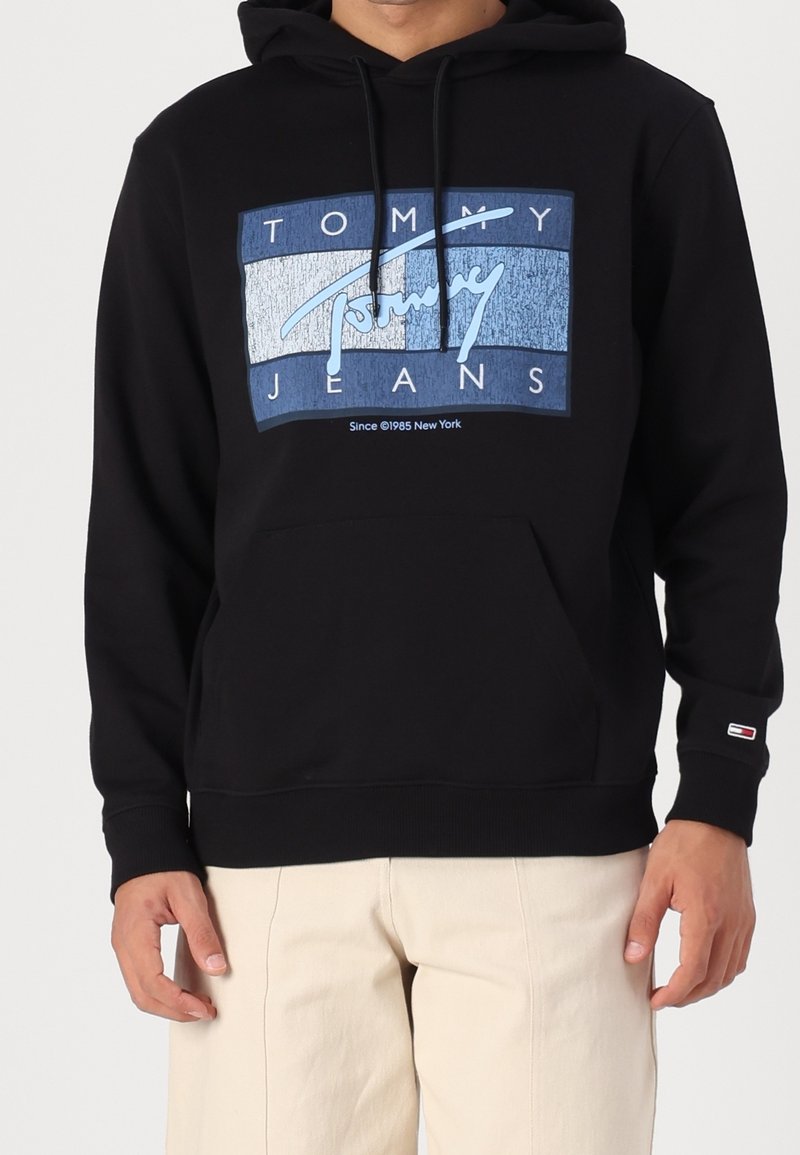 Sweat à capuche noir avec logo Tommy Jeans bleu et blanc sur la poitrine, porté par une personne avec un pantalon beige clair, vu de face du cou jusqu’aux hanches.